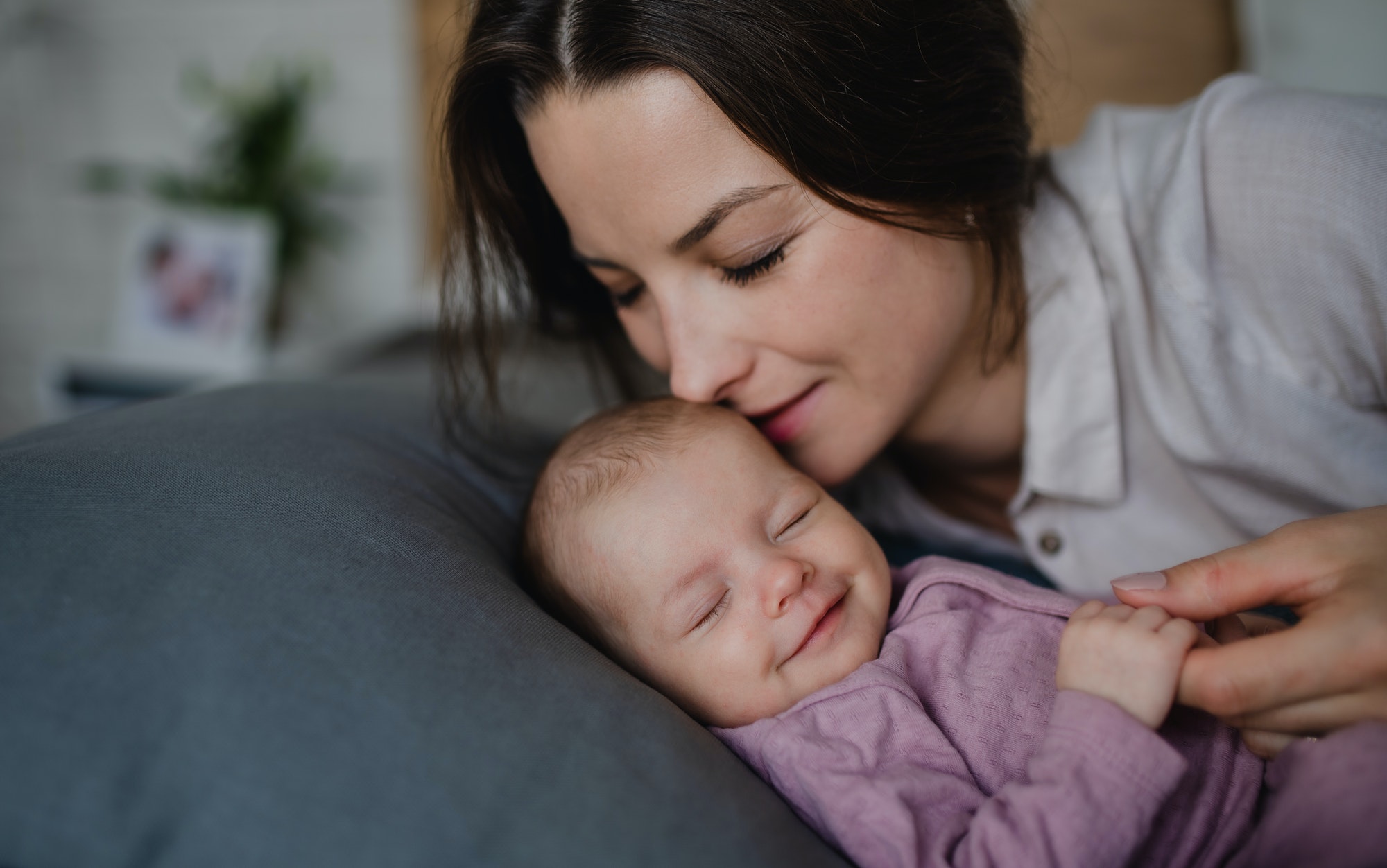 Happy young mother kissing her newborn baby girl, lying on sofa indoors at home